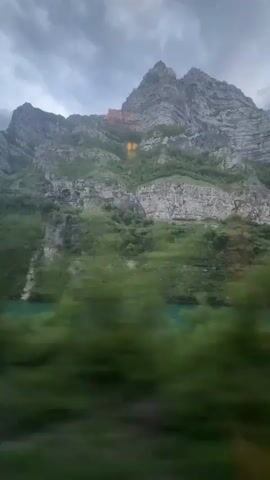 The video shows a view of a tall, rocky mountain against a cloudy sky. The mountain is partially covered in green vegetation. There are two vertical orange streaks on the side of the mountain. The camera is moving, and green vegetation is visible at the bottom of the frame.