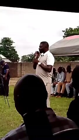 A man in a light-colored collared shirt and pants holds a microphone and speaks. He is standing on a grassy area. Behind him, a group of people are seated, and beyond them is a fence and some trees. A tent is partially visible in the upper right corner. The lighting suggests it is daytime.