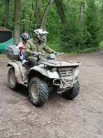 A person wearing a helmet and a green jacket is driving a four-wheeled ATV. A child, also wearing a helmet and a pink shirt, is sitting behind the driver on the ATV. The ATV has large tires and a cargo rack on the back. The ATV is on a dirt path in a wooded area. There are trees on either side of the path, and a building with a dark roof is visible in the background, along with a green tarp covering something. The overall scene suggests an outdoor recreational activity.