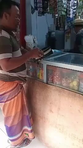 A man wearing a striped shirt and a sarong stands at a counter. He is holding a wallet and appears to be making a transaction. Behind him, a display case filled with various items is visible, and another person can be seen in the background. The environment appears to be a small shop or market stall.