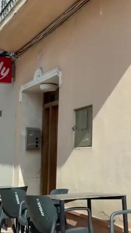 The video begins by showing a storefront with a red and white sign that reads "illy". In front of the store are tables and chairs. The camera then pans to the right, revealing a closed metal shutter. Next, the camera shifts to show a street scene with a building that has pink walls and yellow shutters. There are tables and chairs set up outside this building. Further down the street, a bridge is visible, with a mountain landscape in the background. The video captures a brief view of the environment from what appears to be inside a vehicle.
