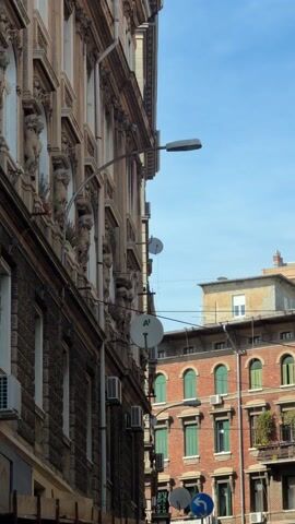 The video shows a view of buildings in a European city. The camera pans upward, revealing detailed architecture with intricate carvings and window shutters. Satellite dishes are visible mounted on the buildings. The sky is clear and blue. The buildings are constructed of stone and brick. There is a street sign visible at the bottom of the frame. There are air conditioning units mounted on the exterior walls of the buildings.
