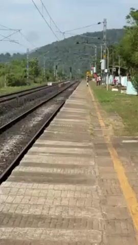 The video shows a train station platform on a sunny day. The platform is made of light-colored concrete blocks with a yellow safety line near the edge. Train tracks run parallel to the platform. In the background, there are green, tree-covered hills. Some people are standing or walking near a building at the far end of the platform. Overhead power lines run along the tracks. The camera pans slowly along the platform.