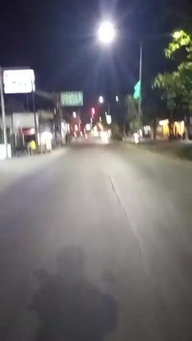The video shows a first-person perspective of someone riding a motorcycle at night on a street lined with buildings and trees. Streetlights illuminate the scene, casting long shadows. Traffic lights are visible, and one appears to be red. A few people are seen walking on the sidewalk. The road is paved, and there are some parked vehicles. The overall atmosphere is that of a quiet, somewhat deserted street at night.