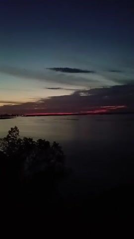 This media file captures a serene sunset over a body of water, likely a river or lake, at dusk. The sky transitions from a deep twilight blue at the top to soft hues of orange and pink along the horizon, indicating the sun has recently set or is about to dip below the landscape. Silhouetted trees in the foreground frame the scene, adding depth and a sense of natural enclosure. In the distance, a faint line of lights suggests a town or settlement on the far shore. The water's surface is calm, reflecting the muted colors of the sky.

The overall mood is tranquil and peaceful, evoking a sense of calm at the end of the day. The time of day is clearly twilight, with the diminishing light casting long shadows and creating a soft, atmospheric effect. There is no visible text in the media file. The setting appears to be a natural landscape overlooking a body of water, consistent with the provided location context of Trypillia, Ukraine. No people or specific objects are discernible in the image, the focus being solely on the natural beauty of the sunset and its surroundings.
