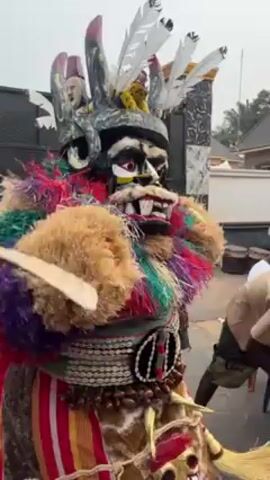 In an outdoor scene, a colorfully dressed African masquerader is seen. The masquerader wears a dark mask with pronounced teeth and large eyes. The costume features a vibrant mix of colors like red, green, purple, and beige, adorned with layers of fringed material, shells, and beadwork. Several white feather-like plumes extend from the top of the mask. People are seen moving around the masquerader. In the background, there's a glimpse of residential architecture.