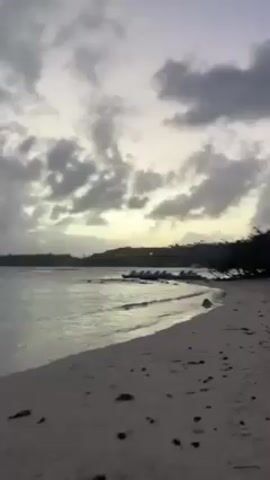 The video shows a beach at dusk or dawn. The sky is filled with grey and white clouds, with a hint of yellow light breaking through on the horizon. The beach is sandy and lined with trees to the right. Gentle waves are lapping at the shore. Several jet skis are parked on a small rocky area near the water. The scene is still and quiet, with no people visible.