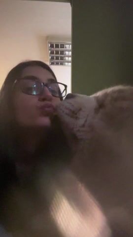 The video shows a person with dark hair and glasses puckering their lips as if to kiss something. They seem to be holding a fluffy, light-colored dog or possibly a large cat. The person then smiles briefly. The background includes a dark green wall, a white ceiling vent, and possibly a doorway. The lighting is somewhat dim.