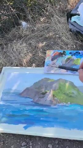 A close-up shot shows a person painting a landscape outdoors. The painting depicts a body of water with waves in the foreground and a green and rocky cliff in the background. The artist is using a brush to add details to the water. A palette with various colors is also visible near the painting. The environment appears to be dry and grassy, suggesting a natural setting. The camera angle is overhead, focusing on the painting and the artist's hand.
