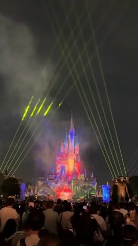 A nighttime outdoor fireworks and laser light show is projected onto a castle, which is illuminated with bright, colorful lights. The castle is tall and ornate, with spires and turrets. Numerous people are gathered in front of the castle, facing it and looking up at the projections and lasers. Many are holding up their phones, presumably to record the event. The sky is dark and appears cloudy. Green lasers are visible emanating from above and pointing towards the castle, with some of them creating a grid-like pattern. The crowd is dense, and the overall atmosphere appears festive and engaging.