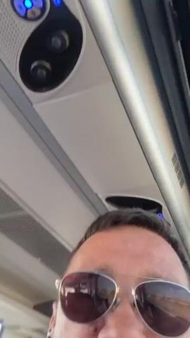 A selfie video is taken from inside a bus. The camera is angled upwards, showing the top interior of the bus with air vents. A man wearing sunglasses is visible at the bottom of the frame. The bus appears to be in motion, with a blurred view of a highway and dry, brown landscape visible through the window. The sky is blue.