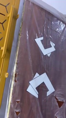 A video showing a close-up of a door wrapped in plastic. The door is made of dark wood and has a repeating pattern of white geometric shapes and thin black lines. There are several tears in the plastic wrap, revealing small triangular sections of the door. A yellow object with a diagonal pattern is visible on the left side of the frame. The lighting is somewhat dim, and the environment appears to be indoors, possibly a storage area or warehouse.