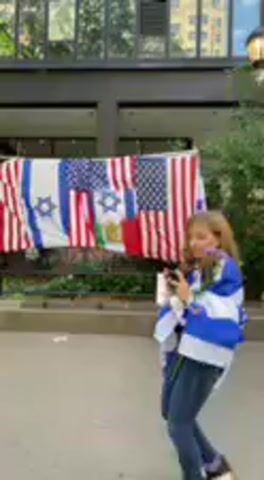 This media file captures an outdoor gathering in what appears to be an urban courtyard or plaza, likely in New York, United States, during the daytime with bright, clear weather.

The main visual focus is a large display of flags hanging horizontally in the background. This display includes multiple American flags, at least two Israeli flags (white with a blue Star of David and blue stripes), and other flags with green, white, and red colors. The flags are suspended in front of a modern building with large glass windows and dark framing.

In the mid-ground and foreground, a group of people is gathered. Sound equipment, including speakers and what seems to be a mixing board, is set up on the left side of the frame, suggesting a planned event or performance. Individuals are seen standing and moving around the area. One person wearing a blue shirt and jeans is visible, and a small, light-colored dog can be seen near some attendees. At the beginning of the video, a young woman wearing a blue and white striped top is seen on the right, looking at her phone.

The setting is characterized by paved ground, surrounding trees, and other buildings in the background. Small, white signs are placed below the flag display, but any text on them is illegible. Throughout the video, Hebrew music plays, setting the auditory tone for the gathering. The overall scene suggests a community event or celebration possibly linked to the national identities represented by the flags.