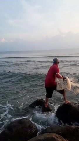 A man stands on rocks in shallow ocean water at a beach and casts a fishing net. He is wearing a red long-sleeved shirt, dark shorts, and a cap.  He casts the net out over the water in a wide arc. The ocean has small waves coming in, and the sky is overcast.
