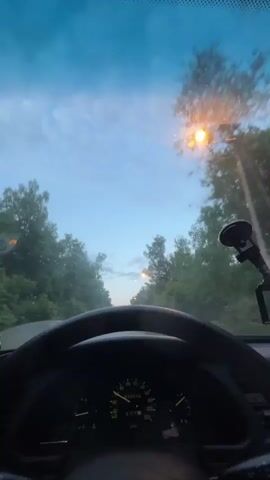 The image is captured from the driver's perspective inside a car, looking out through the windshield. The upper portion of a dark steering wheel is visible at the bottom of the frame, along with the dashboard instruments.

The dashboard features a speedometer, which shows a reading of approximately 20 km/h and is marked up to 220 km/h. To its left, a tachometer displays RPMs up to 7 (x1000). A fuel gauge and temperature gauge are also visible. The odometer displays a trip reading of "000840" and a total mileage of "001844." Several warning lights, including a battery icon and an oil pressure icon, are illuminated.

To the right, a black suction-cup phone mount is attached to the windshield.

Beyond the windshield, a straight, two-lane road stretches into the distance, flanked by dense green trees on both sides. The sky overhead is a soft blue with scattered light clouds, indicating dusk or early evening. A brightly lit street light is visible amongst the trees on the right side of the road, casting an orange glow.

The scene depicts a tranquil drive in a rural or semi-rural area, identified as Bogdanikha, Russia. No other people are visible, and the overall atmosphere is calm.
