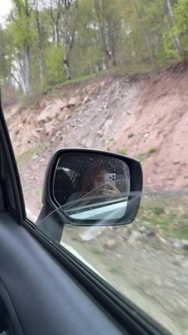 The video shows the driver's side mirror of a car moving along a road with a hillside covered with trees and rocks visible outside. In the reflection of the side mirror, a person, possibly the driver, is visible holding a phone and taking a picture or video of themselves. The mirror has a crack in it. The landscape outside the window appears to be a rural or mountainous area.