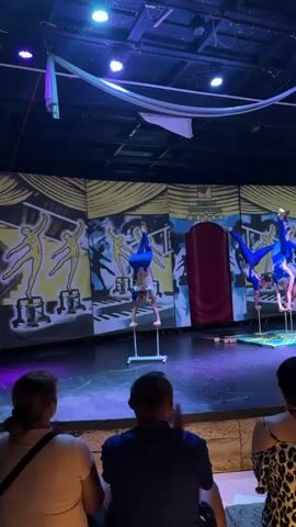 Three people are performing an acrobatic routine on a stage in front of a colorful backdrop depicting stylized figures and musical notes. The performers are wearing blue, form-fitting costumes. Two performers are balancing on their hands on individual stands, while a third performer is balanced on top of them. Another performer is on the right side of the stage, also balancing on their hands on a stand. In the foreground, the backs of three audience members are visible. The audience members are seated on a raised platform. The stage is lit with blue and purple lights.