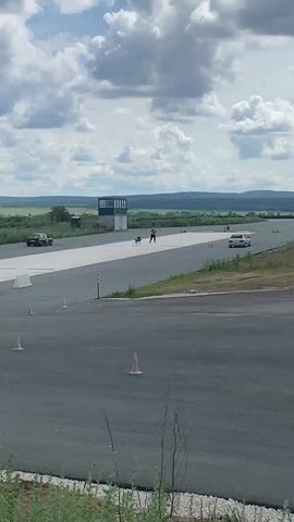 A drone is visible in the foreground, suspended in the air above an asphalt surface. In the midground, a person in an orange shirt and black shorts stands on the asphalt, holding a drone controller. A black car is parked to the left of the person, and a white car is driving on the asphalt to the right. A control tower is visible in the background, with a trailer attached to it. The sky is cloudy, and rolling hills are visible in the distance. The drone appears to be actively flying and controlled by the person in the orange shirt.