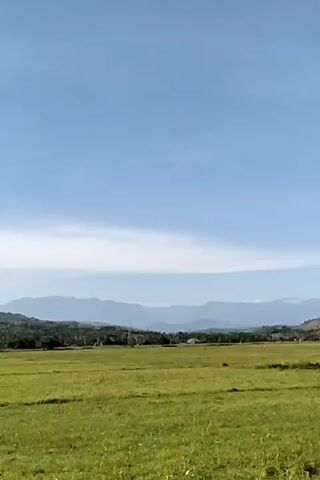 The video shows a green field with mountains in the background under a mostly clear blue sky. The field appears to be grassland or pasture. A line of trees and some structures can be seen between the field and the mountains. An object briefly appears to fly through the frame from left to right, leaving a sound effect, possibly indicating a jet or plane. The scene is static for the duration of the video.
