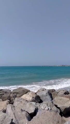 The video depicts a rocky coastline with the ocean visible beyond.  The foreground is dominated by large, grey rocks.  The ocean is a turquoise color, with small waves breaking along the shoreline.  The sky is a clear, pale blue. There is a strong wind blowing.