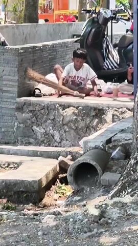 A young male sits cross-legged on a mat outdoors, holding a broom made of natural fibers. He is wearing a white t-shirt with red and black lettering and patterned shorts. To his right are two cups and a plastic bag. Behind him is a dark-colored scooter. The background shows a stone wall and trees, with people visible further in the distance. A concrete pipe is prominent in the foreground.