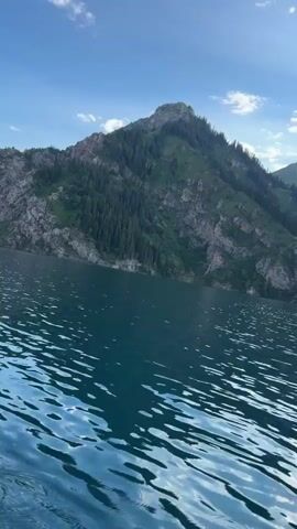 The video shows a wide-angle view of a body of water with mountains in the background. The water is a deep blue-green and has ripples on the surface, indicating movement, possibly from a boat. The mountains are steep and rocky, covered in patches of green vegetation and trees, primarily on the slopes. The sky above is blue with scattered white clouds, and the sun is visible, casting light on the scene. The camera appears to be moving slightly, possibly from a boat on the water, creating a dynamic perspective of the landscape.
