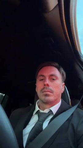 The video is a close-up, front-facing selfie video of a man sitting in the driver's seat of a car. He is wearing a black suit jacket, a white dress shirt, and a patterned tie. The seatbelt is fastened across his chest. The background is dark, suggesting the interior of a car, with a hint of the sky visible at the top right of the frame. The man is looking directly at the camera with a neutral expression. The lighting casts a reddish hue on his face and the left side of his head. There are subtle shifts in his facial expression and head position throughout the brief video, but no significant actions or interactions are depicted.