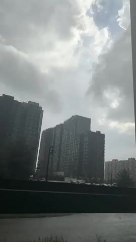 The video shows a view from a window or balcony, looking out at a street with tall apartment buildings. It appears to be raining, as the ground is wet and there is a person walking with an umbrella. Several cars are parked in a lot in front of the buildings. The sky is cloudy and overcast. The camera is static throughout the short clip.