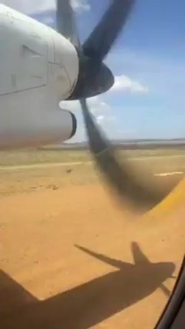 The view from the window of a small aircraft shows the plane on the ground, with its propeller spinning. The ground is red dirt, and in the distance, the terrain is flat with sparse vegetation. The sky is blue with scattered clouds. The shadow of the aircraft's wing is visible on the ground below the window.