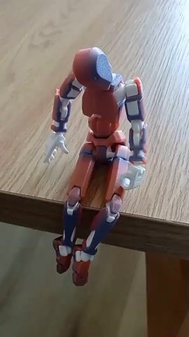This video shows a close-up, slightly angled, top-down view of a red and blue articulated robot figure. The figure is sitting on the edge of a wooden table, with its legs hanging over the side. The robot has white hands, and its body is segmented with visible joints. The lighting suggests it is daytime, with natural light illuminating the scene. The background consists of the wooden table surface and hints of the floor below. The robot is stationary throughout the clip.