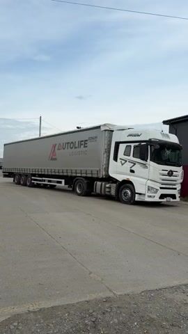 A white FAW Eagle semi-truck with a grey curtain-sided trailer is shown in an outdoor setting. The truck is either stationary or moving very slowly, as the camera pans along its side from front to back, then zooms in on the cab. The word "FAW" is prominently displayed on the truck's grille and above the windshield, with "EAGLE" written below the grille. The trailer has "AUTOLIFE LOGISTIC" and "BONUM MAIN LINE" branding on its side. A Russian license plate, "Q 462 AP 196," is visible on the front of the truck. The sky is overcast, and the ground is paved. A building is visible in the background. A decorative item hangs from the rearview mirror inside the truck's cab.
