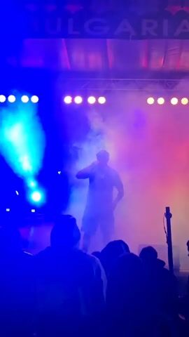 The video shows a rapper performing on a stage filled with colored lights and smoke. The rapper is wearing shorts and a t-shirt and a backwards baseball cap and holding a microphone.  He is the only person on stage, but the silhouettes of audience members are visible in the foreground. The background features a backdrop with the words "SOUND KYHOB$" repeatedly printed on it. The lighting is a mixture of blue, purple, red, and green. The video appears to have been taken by someone in the crowd.