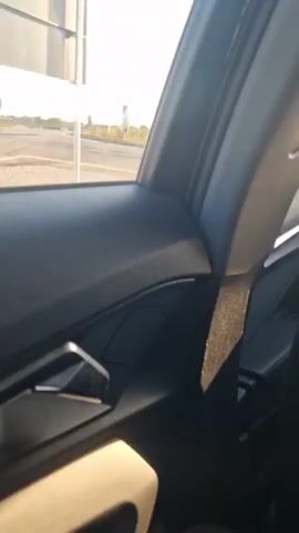 The video captures a view from inside a vehicle, looking out the passenger window. The scene outside shows a road with a curve, bordered by dry grass and some bushes. There are traffic signs visible, including a sign with red and white chevrons indicating a curve and a larger sign with text that is unreadable due to glare. The sun is shining brightly, creating lens flare and casting shadows. The road appears to be paved, with white lane markings. In the distance, there are some structures and trees, suggesting a rural or semi-rural environment. The sky is clear and blue. The perspective is from a moving vehicle, as evidenced by the slight blur and the changing scenery.
