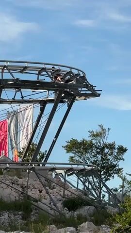 A person is riding a roller coaster cart down a track. The cart is on a metal track suspended above a rocky, grassy slope. There are flags hanging from a structure to the left of the track. The sky is blue with some clouds.