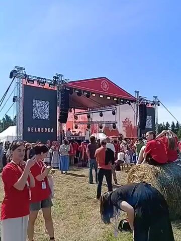 This is a wide-angle video showing an outdoor festival. A large stage with a red roof is set up with a black screen displaying the word "BELOBOKA" and a QR code. There are speakers and lighting equipment above the stage. A crowd of people, some dressed in traditional clothing, are gathered in front of the stage. Several large hay bales are scattered in the foreground and mid-ground, with some people sitting on them. In the foreground, two women in red shirts are visible, one of whom is holding up a phone, possibly taking a picture or video. A person with blue-dyed hair is bending over in the lower right corner. In the background, more people are visible, and the area appears to be a grassy field with trees in the distance under a clear blue sky. A person with a camera on a tripod is standing on a hay bale on the right side of the frame. The overall atmosphere appears to be festive and sunny.