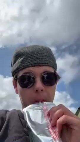 Okay, here's a detailed description of the video:

The video shows a person taking a drink from a foil pouch with a straw. The person is wearing a gray flat cap, sunglasses, and a dark gray t-shirt. They appear to be outdoors, as a blue sky with white clouds is visible in the background. They are looking directly at the camera while drinking. The video then loses focus as the pouch swings out of view, before returning to the subject as they pout at the camera.  The pouch is silver with red markings near the straw.