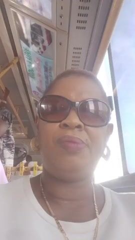 The video features a close-up, selfie-style shot of a woman seated inside what appears to be a public transport vehicle, likely in Kawo, Nigeria. She has short, light brown hair and is wearing large, dark sunglasses, a white shirt, a gold-colored chain necklace, and large hoop earrings. Her facial expressions vary from a slight pout to a subtle smile.

The setting shows the interior of a vehicle, with overhead panels, air vents, and bright daylight visible through the windows, indicating it is daytime. An overhead display or advertisement is partially visible, showing the text "MADA LINE" along with some blurred images and additional text. The background audio includes ambient sounds, suggesting the presence of other passengers and the movement of the vehicle. The woman appears relaxed, looking directly at the camera.