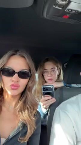 Three young adults are visible from the chest up inside a car. A woman with long, wavy brown hair and black sunglasses sits in the front passenger seat, making hand gestures. A woman with brown hair and glasses sits behind her in the middle, holding a black phone. A man wearing a white shirt sits in the driver's seat, partially visible. The car's interior is dark, with buttons and compartments on the ceiling. The woman in the sunglasses sticks her tongue out and makes a "rock on" hand gesture. The woman in the back smiles and laughs. The man makes a peace sign with his hand.