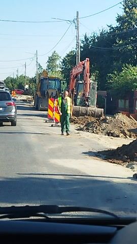 The video captures a road construction scene in Săbăreni, Romania, likely during daytime given the bright sunlight and clear sky. The camera appears to be positioned inside a vehicle, looking out at the road.

The primary subjects are construction equipment and vehicles. An orange excavator is prominent in the foreground, with a worker in a high-visibility vest standing nearby. Further down the road, a yellow bulldozer is also visible, partially obscured by the excavation site. Several cars are on the road, forming a queue, suggesting traffic is being managed due to the construction. These include a dark SUV with a roof rack, a yellow car, and a blue car. Construction barriers with red and yellow diagonal stripes are placed to delineate the work area.

The scene depicts ongoing roadworks. The ground is dug up, revealing earth and possibly underground utilities. Trees and foliage line the side of the road, indicating a semi-rural or suburban setting. Power lines and poles are also visible, typical of roadside infrastructure. The overall impression is one of routine road maintenance or repair.

The time of day appears to be midday, judging by the strong shadows cast by the vehicles and the bright illumination of the scene. The weather seems to be fair and sunny. There is no discernible text in the video, except for what might be on the license plates of the cars, which is not legible. The atmosphere is functional, focused on the construction activity and the flow of traffic.