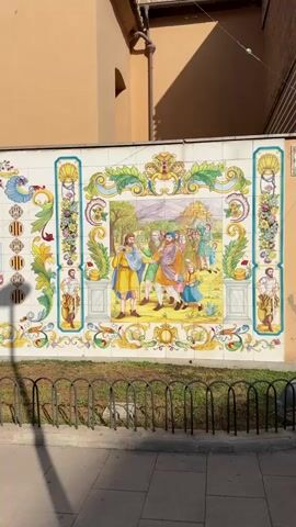 The video shows a tiled mural on the exterior of a building. The mural is colorful and depicts various scenes and emblems. The building appears to be a church with a bell tower visible in the background. The mural is adorned with floral motifs, figures in historical attire, and coat of arms. The environment appears to be outdoors, with a walkway and a fence in the foreground. The lighting suggests it is daytime.