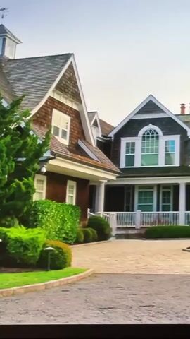 This video displays a large, multi-story house with a circular turret on the right side and a steeply pitched gable roof on the left. The exterior of the house is a mix of dark shingles and lighter siding, with white trim around the windows and a white porch railing. The driveway in front of the house is paved with gravel. There are manicured bushes and trees surrounding the property. The sky above is a pale blue with some clouds, and the overall lighting suggests it is daytime, possibly late afternoon or early morning due to the soft light. The camera appears to be moving, panning across the front of the house.