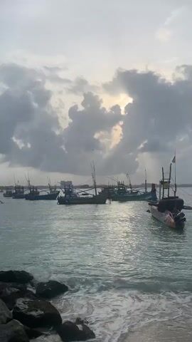 The video shows a body of water with several small fishing boats anchored. The sky is cloudy, with the sun peeking through in the upper right corner. The water is choppy and appears to be near a shore, as evidenced by rocks visible in the foreground. The boats are diverse in color and design, with tall poles, likely for sails or fishing equipment, extending upwards. Some buildings or structures are visible in the distance on the horizon. There are no people visible on the boats or in the water. The overall atmosphere is somewhat overcast and the lighting suggests it might be early morning or late afternoon.