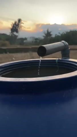 The video shows a close-up, low-angle shot of water dripping from a pipe into a large blue plastic tank. The pipe is made of PVC and is attached to an elbow joint. The tank is partially full of water, and the droplets are creating ripples on the surface. In the background, the sun is setting, casting a warm glow over a rural landscape with trees and fields. The sky is filled with soft colors, and the overall atmosphere is peaceful.
