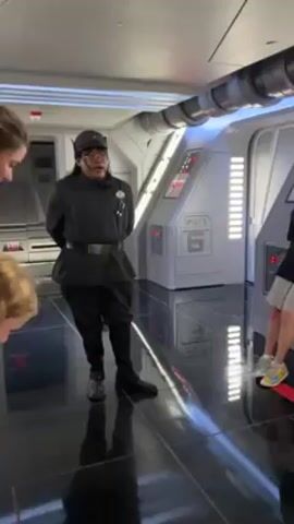 A medium shot shows a person dressed in a Star Wars-like First Order uniform standing in a white-paneled hallway. To the right, there are control panels set into the wall. The hallway's floor is black and shiny. To the left, there is a door with an "exit" sign over it. The First Order employee is speaking and gesturing to a couple of children nearby. They are wearing a black cap with their uniform.