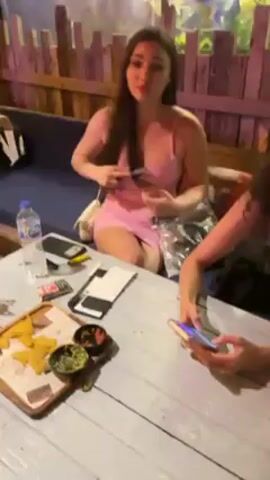 Two women sit at a weathered white wooden table in an outdoor setting with wooden fencing visible behind them. The woman on the left, with long brown hair and wearing a light pink dress, fiddles with a small black object. To her right, the other woman, with curly hair, looks at her phone. On the table, there's a tray with tortilla chips and dips, a water bottle, phone cases, and other items. The atmosphere seems casual and relaxed.