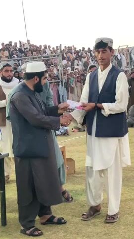 The video shows a group of men gathered outdoors, seemingly at a sports event or ceremony. In the foreground, two men are shaking hands. One man, on the left, is wearing a dark grey tunic, a navy vest, and a white kufi. He is handing a piece of paper to the man on his right. The man on the right is wearing a white tunic, a navy vest, and a white patterned hat. He is receiving the paper. There are trophies on a table to the left of the man in the grey tunic. In the background, a large crowd of people are seated and standing behind a fence, watching the proceedings. The environment appears to be a grassy field with a large open space. The sky is visible in the background, suggesting it is daytime. The overall atmosphere seems formal and celebratory.