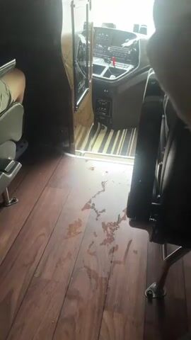 The video appears to be filmed from inside a bus, looking towards the driver's cabin and the road ahead. The floor of the bus is made of dark wood-like material and has several dark wet spots on it. In the driver's cabin, the dashboard is visible, as well as the steering wheel and the driver's seat, though the driver is not clearly visible. There is a large black screen mounted above the dashboard. The bus is driving through a tunnel, and traffic can be seen ahead. The weather appears to be rainy, as evidenced by the wetness on the road outside the tunnel. There are people in the bus, but they are mostly out of focus and partially visible in the foreground.