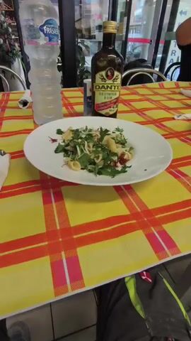 The video shows a table in what appears to be a restaurant or cafe setting, with a bright yellow and orange plaid tablecloth. On the table, there are plates of food, glasses, a bottle of olive oil, and a bottle of water. A person is visible in the background, wearing a black t-shirt. The camera is positioned to show a close-up of the table and the food.

Specifically, there are two plates of pasta with greens on them. One plate is closer to the camera and is in focus, showing a generous portion of pasta with herbs and some red elements, possibly dried cranberries or pomegranate seeds. To the left of this plate, a bottle of "DANTE OLIO EXTRAVERGINE DI OLIVA" is prominently displayed. Next to the olive oil bottle is a shaker of salt. A bottle of "FRASASSI" water is also on the table. There are at least three glasses on the table. A white napkin, folded and placed in a holder, is on the left side of the frame.

In the background, a person in a black t-shirt is partially visible, seated at the table and reaching for something. Another person is faintly visible in the far background, walking away. The setting appears to be indoors, possibly a casual eatery, with what looks like large windows or glass doors in the background.

The lighting suggests it could be daytime, perhaps lunch or early dinner. There are no obvious indicators of the weather. The overall mood seems relaxed and casual, consistent with a meal being served. The visible text includes "DANTE OLIO EXTRAVERGINE DI OLIVA" on the olive oil bottle and "FRASASSI" on the water bottle. The presence of Italian text on the olive oil suggests the location is likely in Italy, which is further supported by the context provided. The image is captured from a slightly low angle, looking across the table.