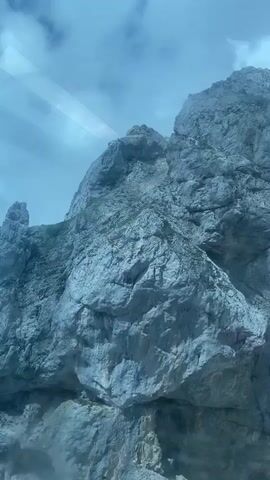 The video captures a view of a rugged, rocky mountain range under a cloudy sky. The camera appears to be moving, offering a dynamic perspective of the steep slopes and jagged peaks. Patches of green vegetation are visible on some of the rocky surfaces, contrasting with the predominantly grey and blue tones of the landscape. Wisps of fog or clouds drift across the mountain faces, adding a sense of depth and atmosphere to the scene. The lighting suggests it is daytime, possibly overcast or at a time of day when sunlight is diffused. There are no people or man-made objects clearly visible, emphasizing the natural and untamed character of the mountain environment.