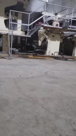 The video depicts a large, stationary industrial machine, likely located inside a factory or warehouse. The machine is primarily beige and black with silver metal guardrails and a set of access stairs. The machine has several visible rollers and mechanical components. The flooring is gray concrete with cracks and some puddles of liquid. The lighting is fluorescent. The camera is static, and there are no people visible in the frame. The environment is utilitarian and industrial.